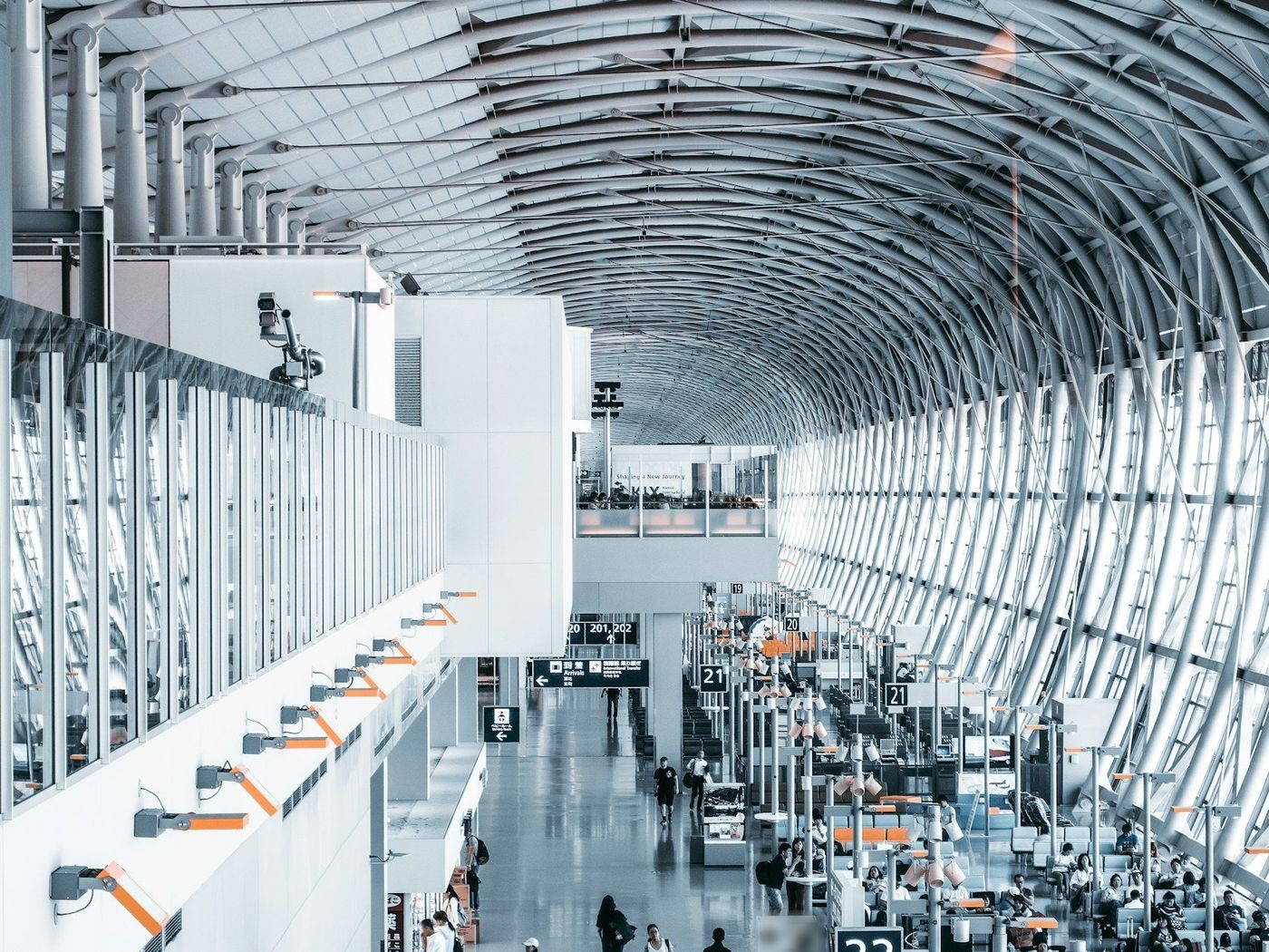 A long airport concourse seen from an upper level, with travelers walking past rows of gate seating under a curved steel roof and glass wall