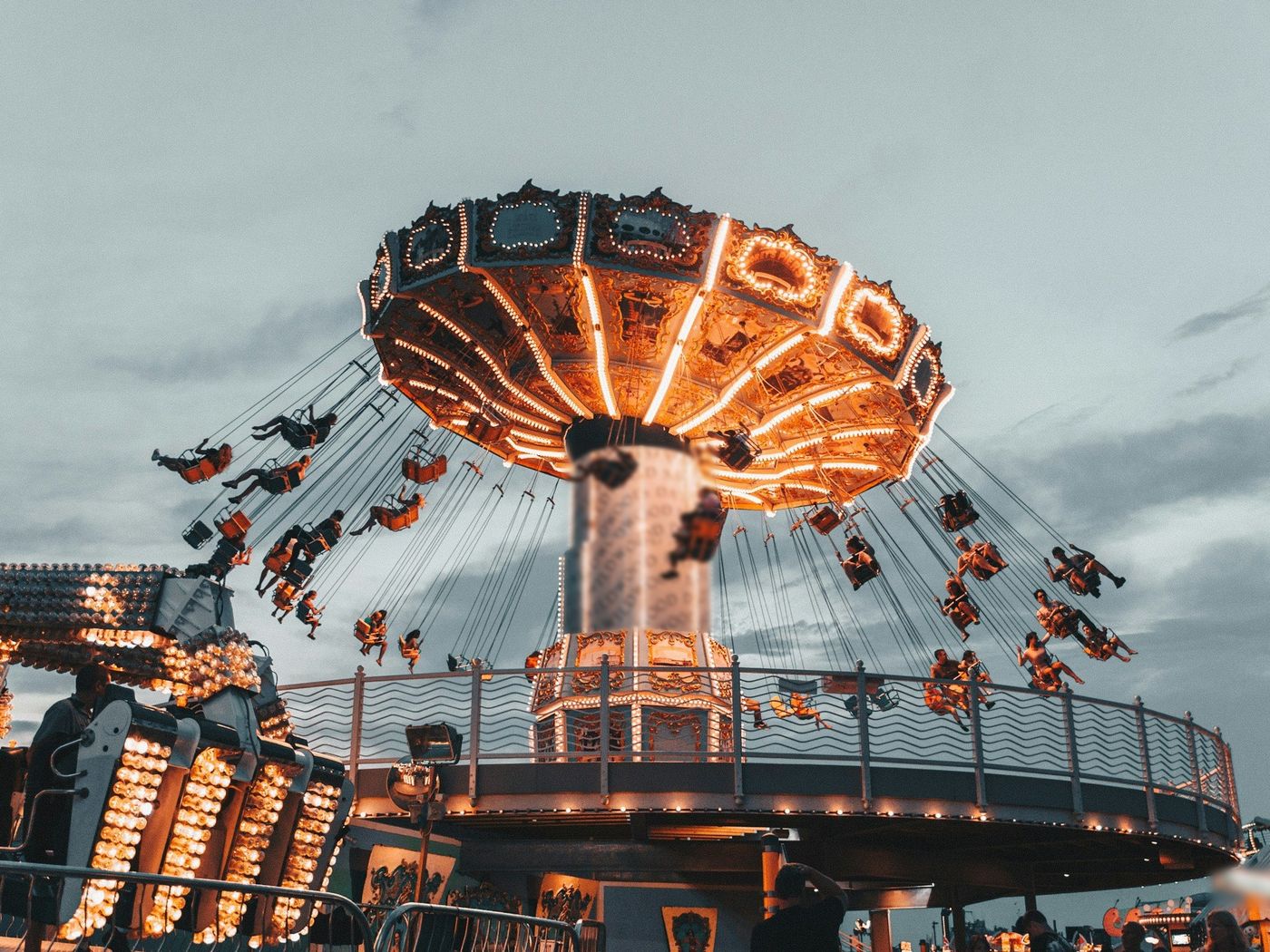A chair-swing ride lit with rows of warm bulbs carrying riders against an overcast evening sky, beside other lit rides and a green coaster track