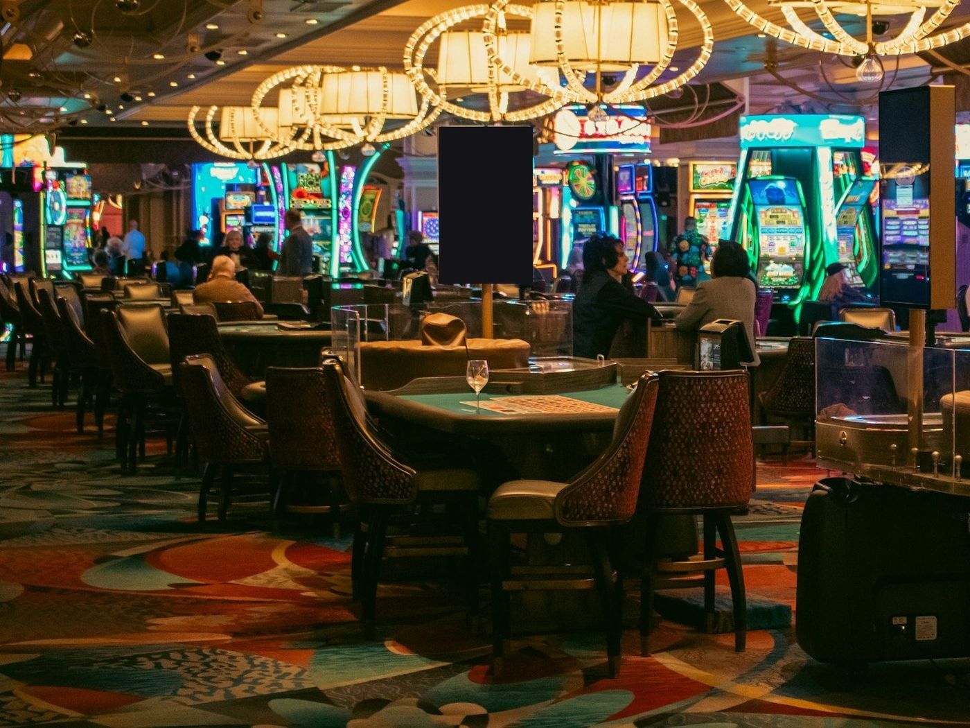 A wine glass on a green-felt gaming table surrounded by empty chairs, with hoop chandeliers overhead and lit slot machines behind