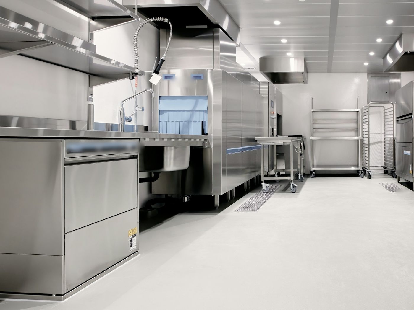 An empty stainless-steel commercial kitchen with a pre-rinse sink, a conveyor dishwasher, wall shelves and rolling carts on a pale gray floor