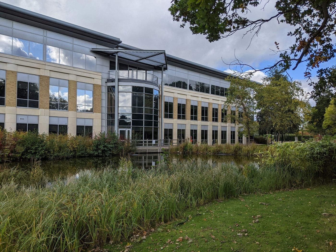 A three-story office building of glass and pale brick beside a reed-lined pond and lawn on a cloudy day
