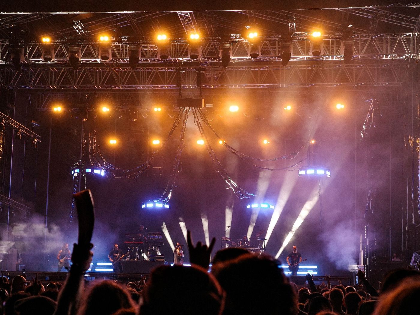A festival main stage at night lit by overhead truss and beams, with a silhouetted audience standing in front