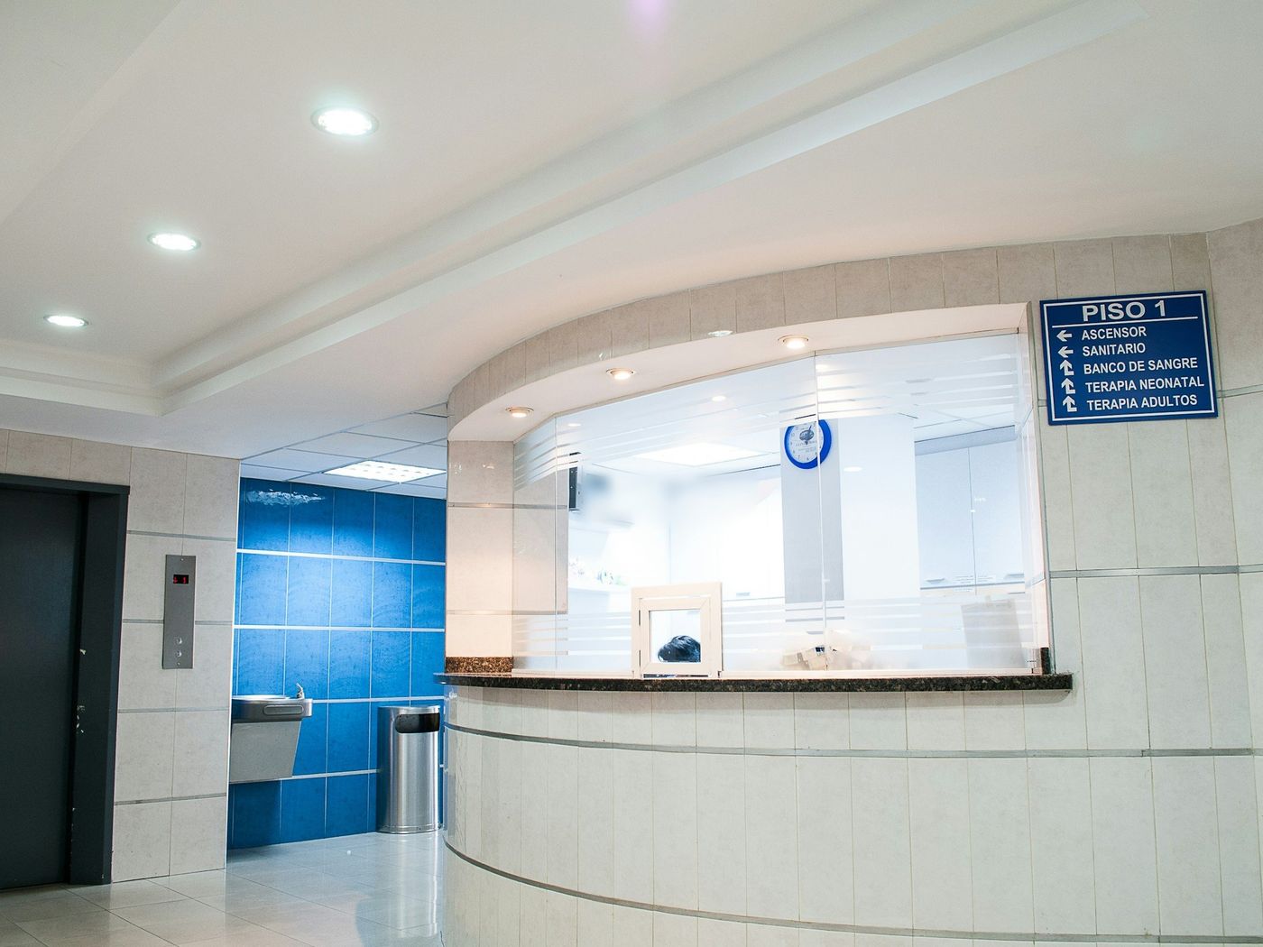 A curved, tiled reception counter with a frosted glass screen in a hospital lobby, beside an elevator, a drinking fountain and a blue direction sign