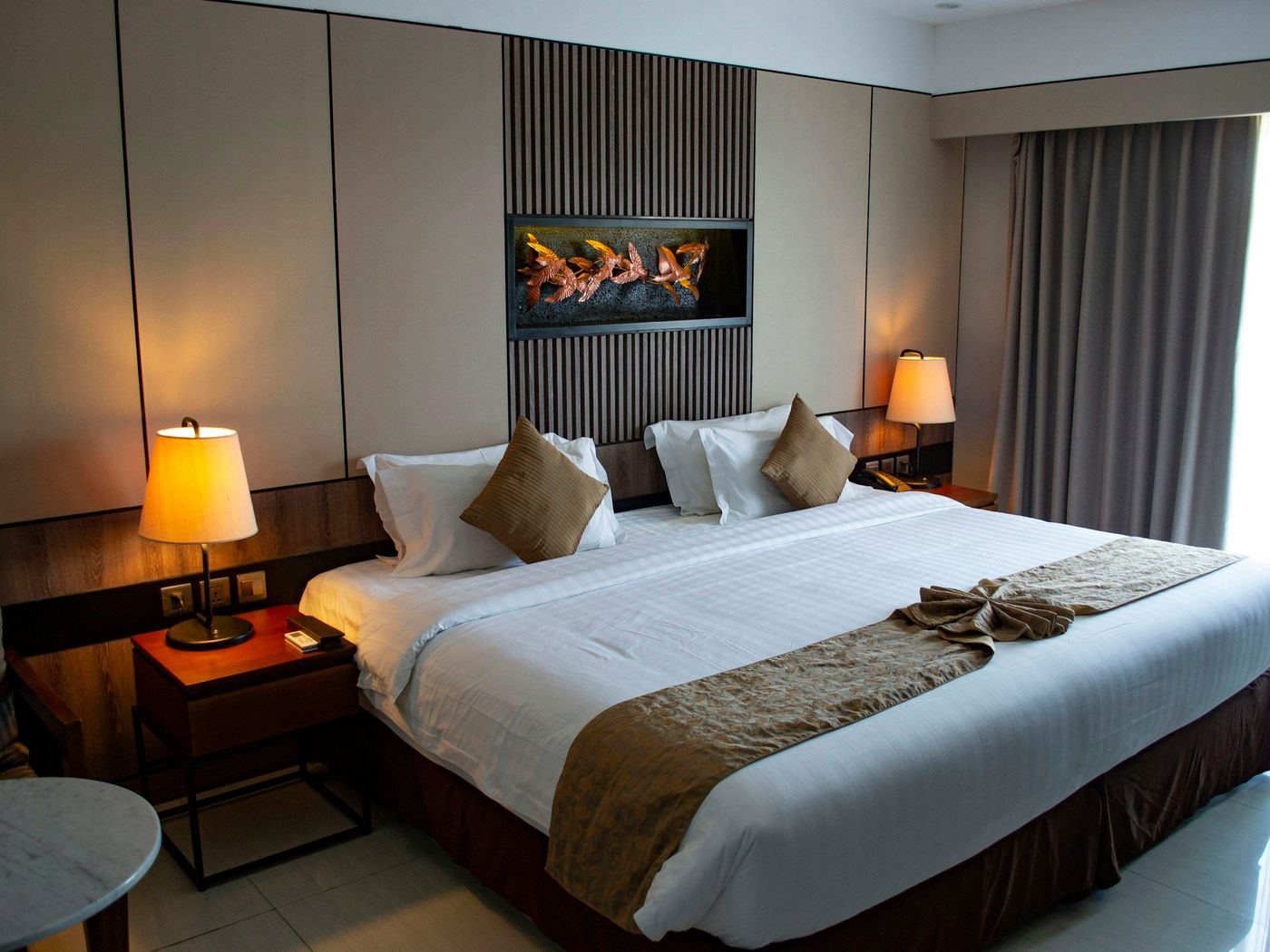 A hotel guest room with a white-linen bed and brown runner, lamps lit on both nightstands and curtains beside a bright window