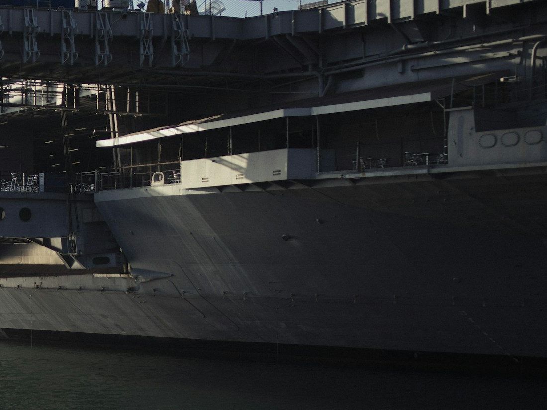 The gray hull of a large naval ship in dark harbor water, with metal tables and chairs set out on a shaded open deck