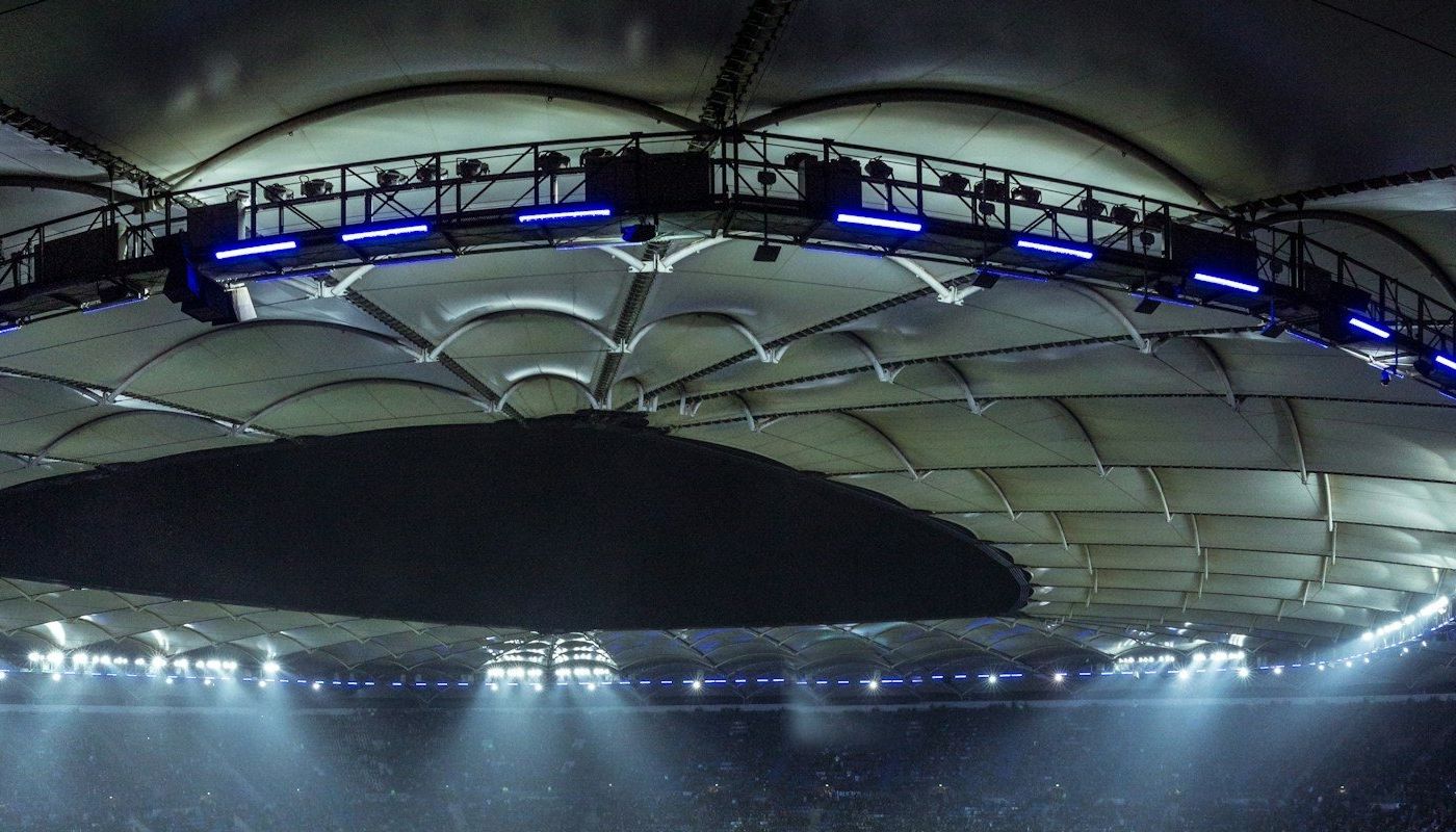 A stadium roof lit by floodlights at night above a crowd in the upper stands