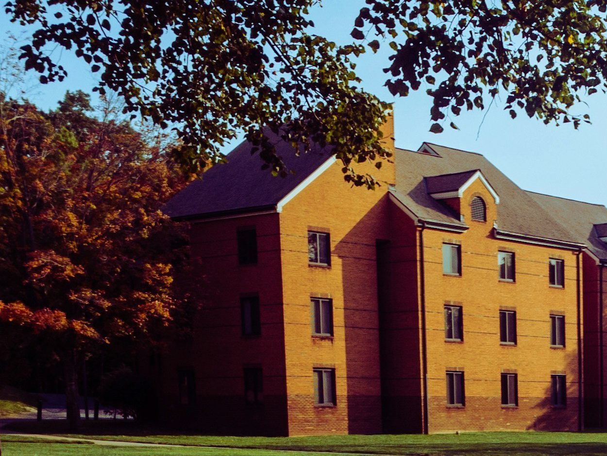 A three-story brick building with a pitched roof in warm, low sunlight, framed by overhanging branches and autumn trees above a lawn